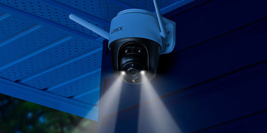 Lorex 2K pan-tilt Wi-Fi security camera mounted under an eave, with built-in dual spotlights illuminating the area at night.