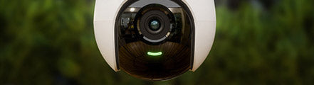 Close-up of a Lorex lightbulb security camera installed outdoors, with a wide-angle lens reflecting the surrounding area and a green indicator light glowing beneath it; blurred greenery is visible in the background.